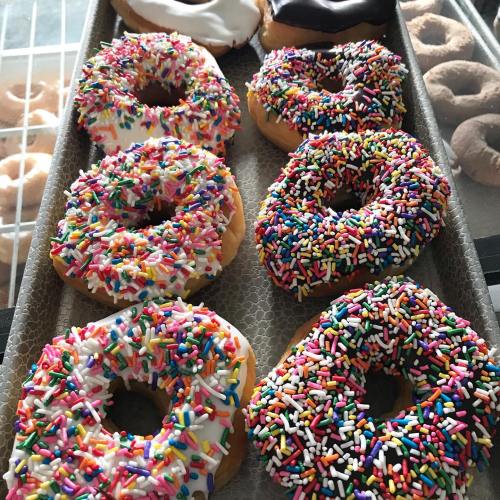 A tray of colorful donuts topped with white icing and rainbow sprinkles, alongside chocolate-covered donuts.