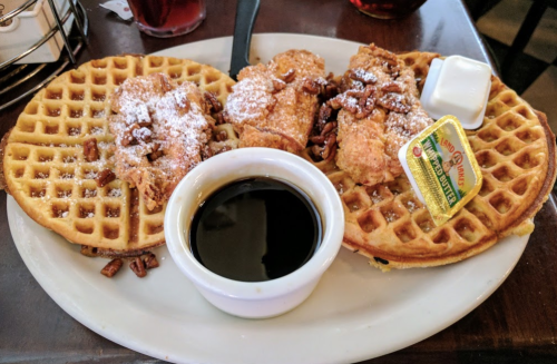 Two waffles topped with powdered sugar and pecans, served with fried chicken, syrup, and a butter pat on the side.