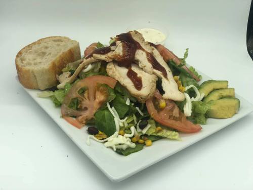 A colorful salad with grilled chicken, tomatoes, avocado, corn, and a slice of bread, served with dressing on the side.