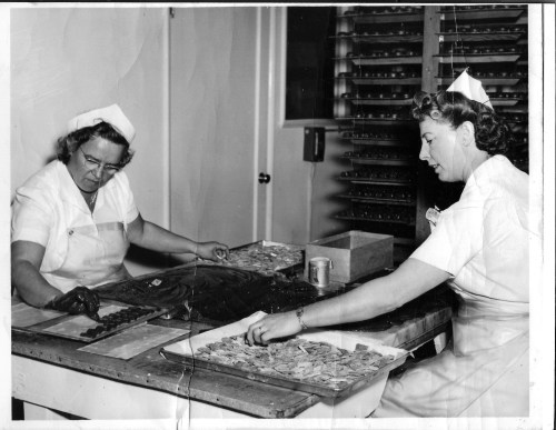 Two women in white uniforms working at a table, sorting and organizing small items in a workshop setting.
