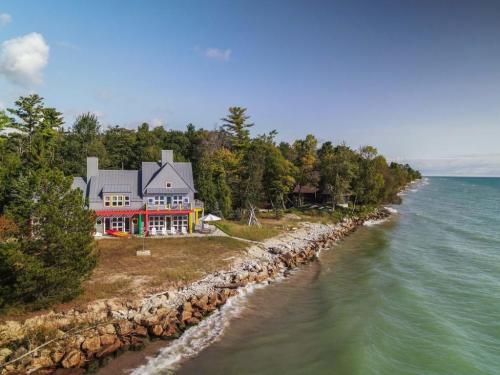 Aerial view of a colorful house by the shore, surrounded by trees and rocky coastline under a clear blue sky.