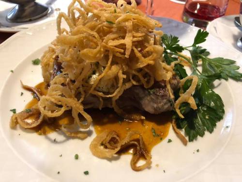 A plated dish featuring a piece of meat topped with crispy onions and garnished with parsley, served with sauce.