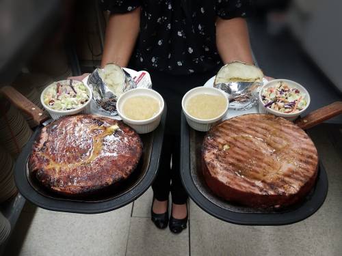 A person holds two trays of grilled meat, baked potatoes, coleslaw, and dipping sauces in a restaurant setting.