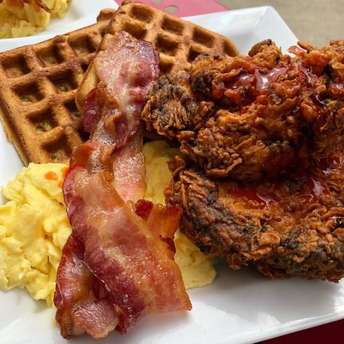 A plate of waffles, fried chicken, scrambled eggs, and crispy bacon drizzled with syrup.