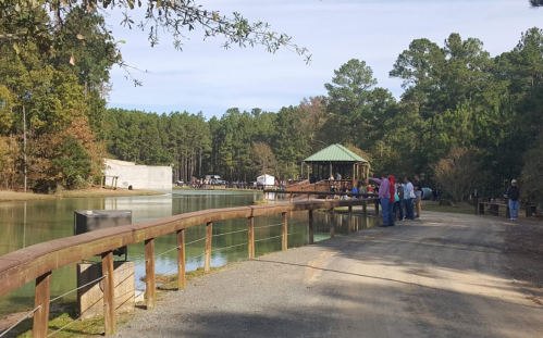 A peaceful lakeside scene with people walking along a path, surrounded by trees and a gazebo in the background.