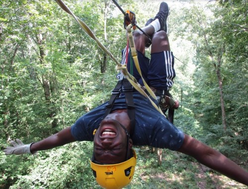 A person in a helmet hangs upside down on a zip line, smiling, surrounded by lush green trees.