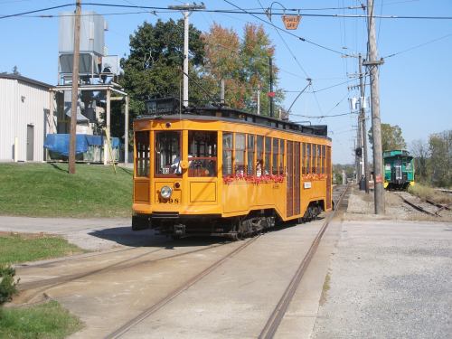 A vintage orange streetcar on tracks, surrounded by greenery and industrial buildings, under a clear blue sky.