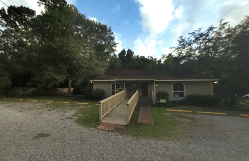 A small, single-story building with a ramp, surrounded by trees and a gravel parking area under a cloudy sky.