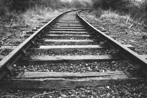 Black and white image of a curved railway track surrounded by overgrown grass and gravel.