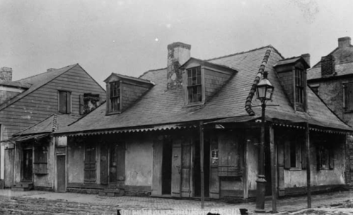 Historic black-and-white photo of a weathered, two-story house with a sloped roof and nearby buildings.