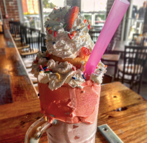 A colorful milkshake topped with whipped cream, sprinkles, and a mini cupcake, served in a glass with a pink straw.