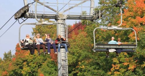 Two chairlifts carry people above colorful autumn trees, with six riders enjoying the scenic view.