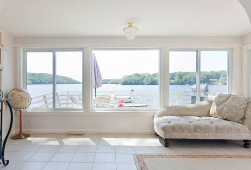 Bright interior with a cozy couch, large windows overlooking a lake, and a small table on a deck outside.