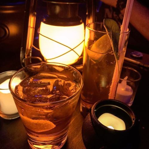 Two cocktails on a table, one with ice and citrus, surrounded by candles and a lantern, creating a cozy atmosphere.