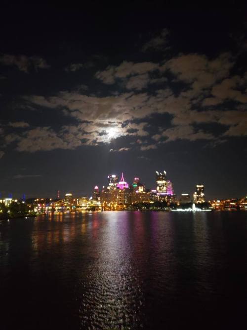City skyline at night with colorful lights reflecting on the water and a bright moon in a partly cloudy sky.