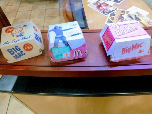 Three Big Mac boxes displayed in a glass case, showcasing different designs and sizes at a McDonald's restaurant.