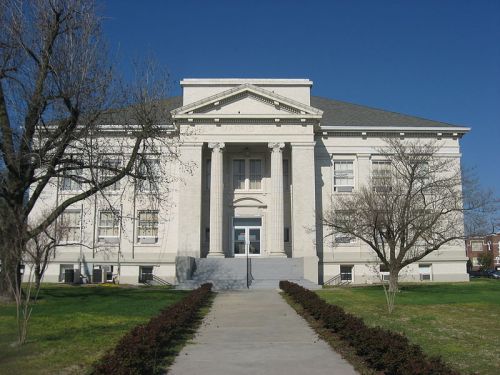 Historic courthouse with a grand entrance, flanked by trees and a clear blue sky. Green lawn in the foreground.