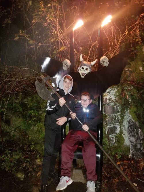 Two boys pose on a spooky throne with skulls and bat wings, holding an axe and a staff, in a dark, eerie setting.