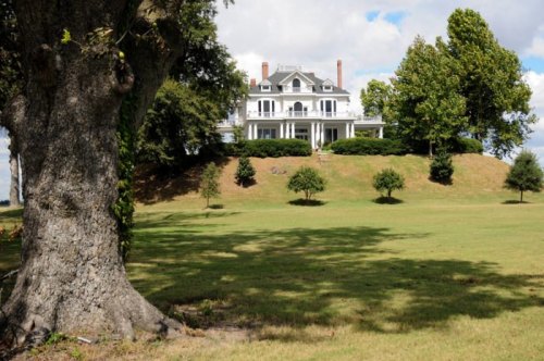 A large white mansion sits atop a grassy hill, surrounded by trees under a partly cloudy sky.