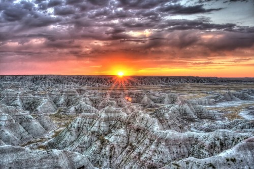 Sunset over rugged, layered rock formations in a vast, open landscape with dramatic clouds.