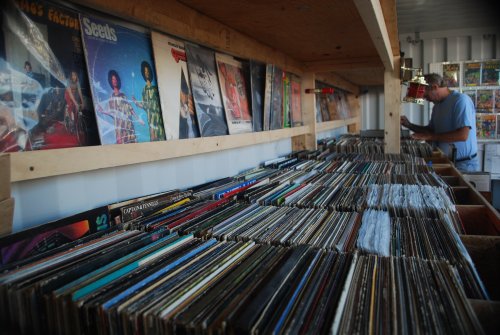 A wooden shelf filled with vinyl records, with a person browsing in the background. Colorful album covers are displayed above.