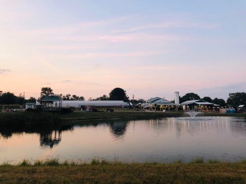 A serene pond reflects a building at sunset, surrounded by trees and a grassy area. A fountain adds to the peaceful scene.