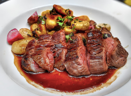 A plate of sliced steak with a rich sauce, served alongside roasted potatoes and garnished with herbs.