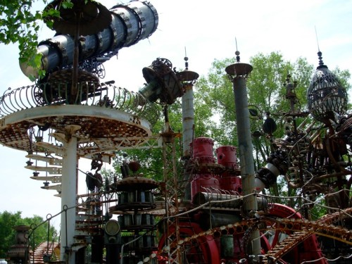 A whimsical sculpture garden featuring large, intricate metal structures and gears surrounded by greenery.