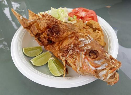 A plate of fried fish served with lime wedges, lettuce, tomato, and a side of mashed beans.