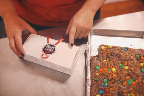 A person points at a white box with a logo, next to a tray of colorful, topped brownies on a metal surface.