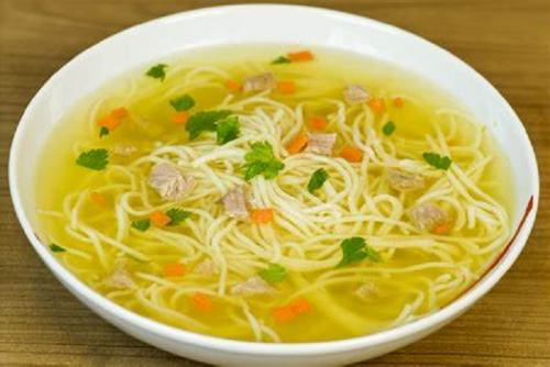 A bowl of clear broth soup with noodles, pieces of meat, and garnished with chopped herbs and diced carrots.