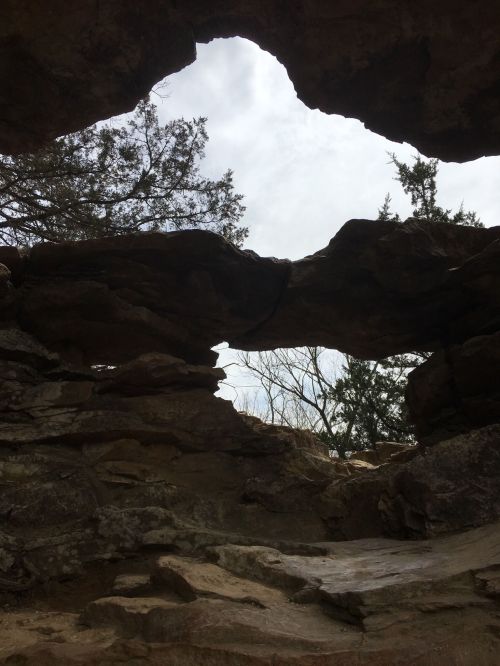 A natural rock formation with a hole, framed by trees and cloudy sky in the background.
