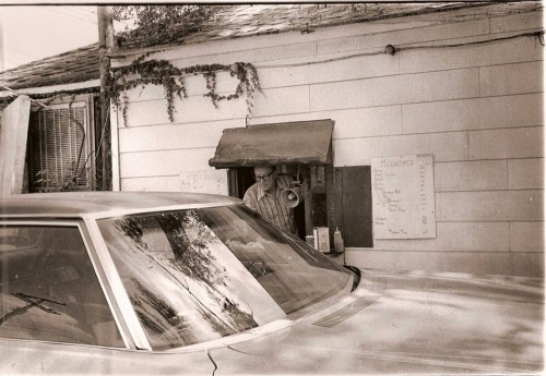 A vintage black-and-white photo of a man at a drive-thru window, with a car parked outside and a menu on the wall.