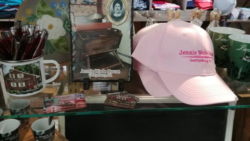 Display of souvenirs featuring a pink cap, mugs, pens, and a photo of the Jennie Wade House in Gettysburg, PA.