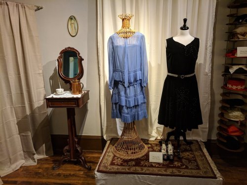 A display featuring a blue dress on a mannequin, a black dress on a stand, and a vintage table with a mirror.