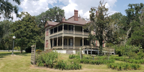 A large, historic house with a wraparound porch, surrounded by lush greenery and trees draped in Spanish moss.