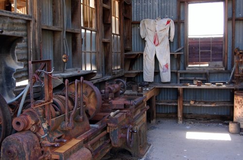 An old workshop with rusty machinery and a white suit stained with red hanging on the wall. Sunlight streams through windows.