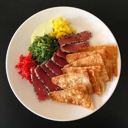 A plate featuring sliced tuna, crispy wontons, seaweed salad, and colorful garnishes.