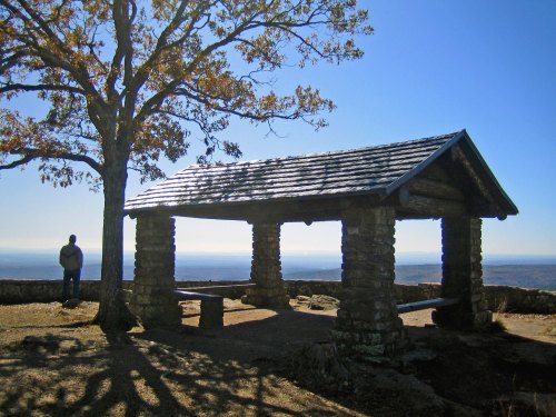 A stone shelter with a wooden roof stands on a hill, with a person nearby and a clear blue sky in the background.