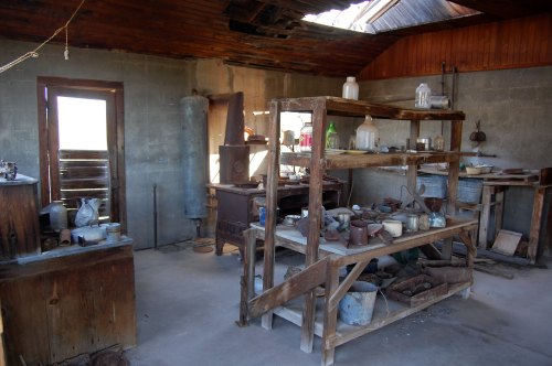 A rustic kitchen with wooden shelves, old utensils, and jars, featuring a stove and a partially open roof.