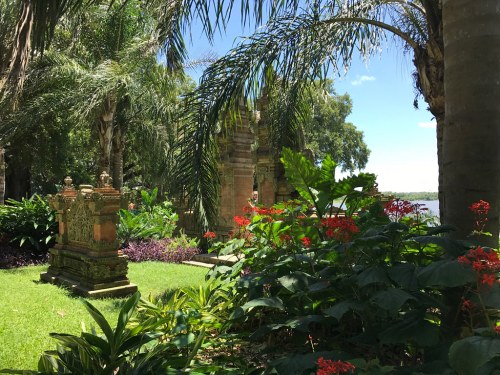Lush garden with tropical plants, vibrant flowers, and ornate stone structures near a body of water under a clear blue sky.