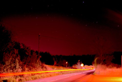A long exposure shot of a dark road at night, with red and orange light trails and stars visible in the sky.
