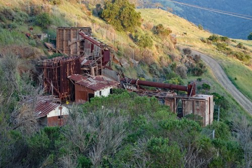 Abandoned industrial structures overgrown with vegetation on a hillside, with a dirt road nearby.