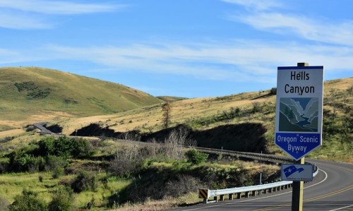 A scenic view of Hells Canyon with rolling hills and a road sign for the Oregon Scenic Byway. Clear blue sky above.