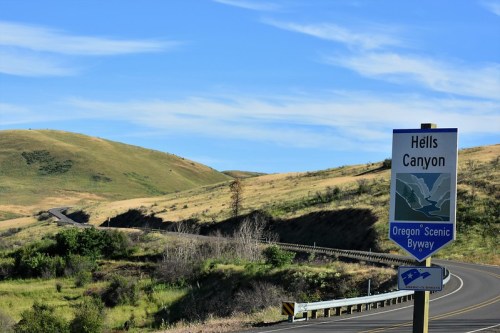 Sign for Hells Canyon along a winding road, with rolling hills and a clear blue sky in the background.