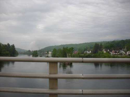 A view of a river surrounded by green hills and houses, seen from a bridge under a cloudy sky.