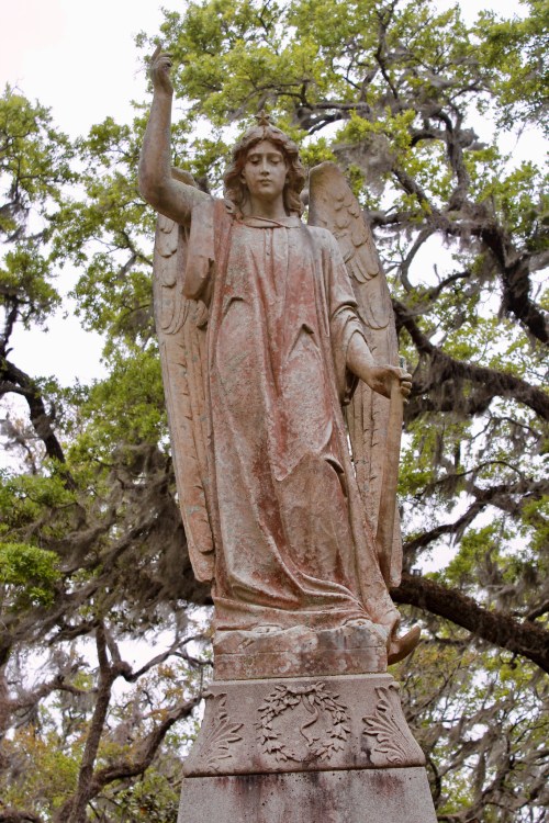 A weathered angel statue with wings raised, standing among green trees and moss.