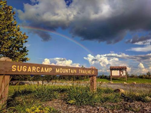 A wooden sign for Sugarcamp Mountain Trailhead with a rainbow and clouds in the background.