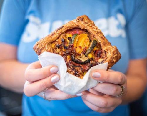 A person holding a flaky pastry filled with colorful vegetables, wrapped in paper, against a blue shirt background.