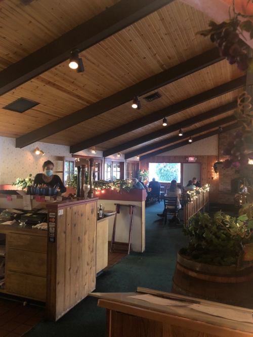 Interior of a cozy restaurant with wooden beams, plants, and patrons dining at tables. Staff member wearing a mask.
